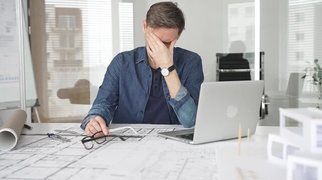 Stressed architect feeling overwhelmed and frustrated, suffering from burnout while working on blueprints for a tight project deadline in a modern office setup