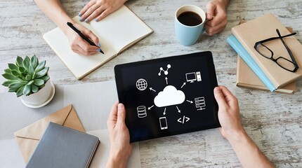 Hands holding tablet displaying cloud computing diagram with icons for globe database phone code computer and servers near notebook coffee and plant technology network internet digital connection