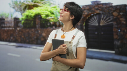 Woman in apron holds tablet to chest while looking up with a smile on a residential street by a stone gate and tree  optimism. © Krakenimages.com