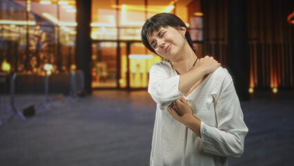 Fototapeta premium Woman holding shoulder and wincing at entrance of a modern building with stanchions and glass doors; pain fatigue.