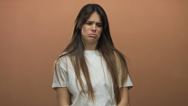 Young hispanic woman with long hair wearing white shirt standing against isolated brown wall background looking sad
