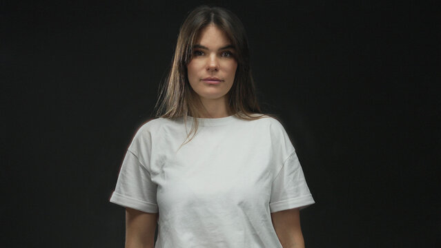 Young hispanic woman making gun gesture with fingers against isolated black background wall, expressing frustration or boredom with casual expression in white shirt.