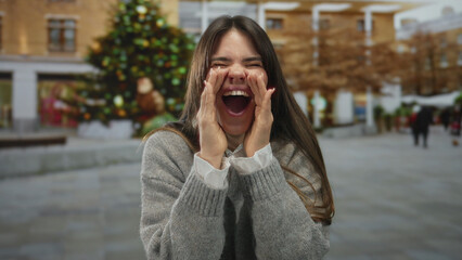 Fototapeta premium Woman shouting joyfully in urban street wearing grey sweater with christmas tree in background capturing festive spirit in city square