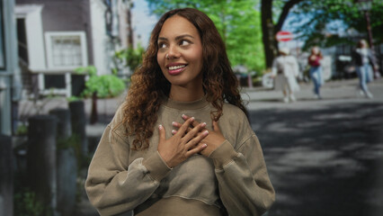 Fototapeta premium Young african american woman smiling with hands over chest on a city street wearing a beige sweatshirt and natural curls; gratitude warmth.