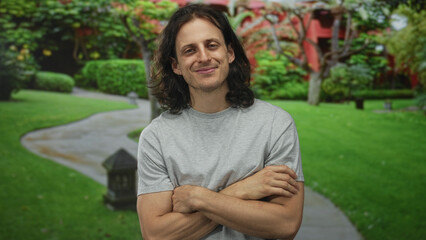 Man with crossed arms showing bare forearm on forest path beside green lawn and stone lantern;...