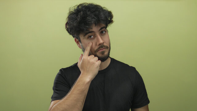 Young hispanic man with beard pointing at his eye against a green background expressing curiosity and contemplation in a studio setting.