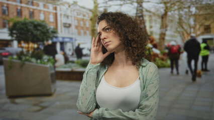 Fototapeta premium Woman touching temple with fingers and crossing arm over chest on city street near planters and trees; thoughtful contemplation.