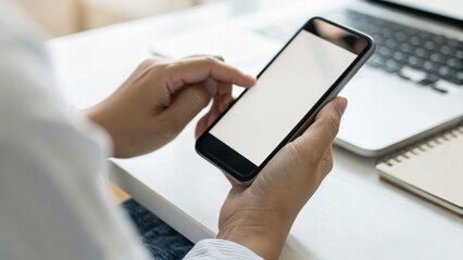 A person using a smartphone while sitting at a desk with a laptop and notebook in the background.