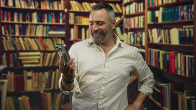 Man holding smartphone in right hand, left hand raised while smiling at the screen in a library building; contentment curiosity learning.