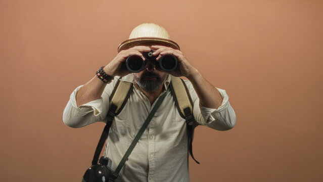 Man wearing pith helmet holding binoculars and looking through them in a peach studio; curiosity travel adventure.