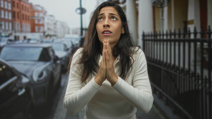 Young hispanic woman praying with hands pressed together in a pleading gesture on a city street...