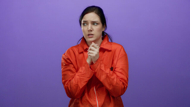 Woman with brunette hair wearing orange prison jumpsuit stands in front of purple background, appearing worried.