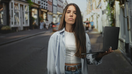 Fototapeta premium Young hispanic woman points finger to camera while holding a laptop and pinching her nose on a busy street; disgust caution.
