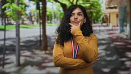 Woman wearing a gold medal, hand on chin and pointing up while standing on street in mustard sweater; pride reflection.