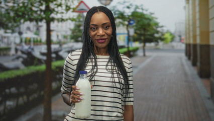 Fototapeta premium Black woman holding milk bottle with hand visible, braided hair and striped top on a city street with blurred buildings and sidewalk; confidence wellness.