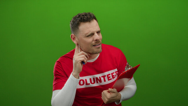 Young man in a red volunteer shirt listens and writes on a clipboard against a green wall, focusing attentively.