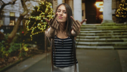 Fototapeta premium Young woman with long hair and braces reaches forward with both hands and outstretched palms on stone steps of building near lit trees at night; defiance rebellion.