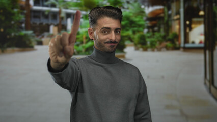Fototapeta premium Man points finger to camera on street lined with potted plants and modern building facades while wearing a gray turtleneck; confidence greeting.