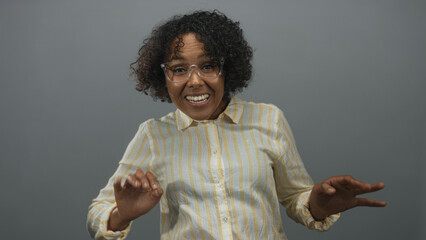 Young african american woman wearing yellow striped shirt smiling waves hands in grey studio;...