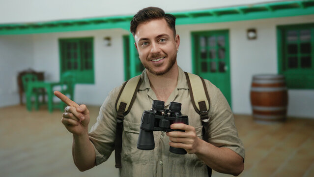 Young tourist man with a beard, holding binoculars and pointing on a city street, wears a backpack, suggesting adventure and exploration.