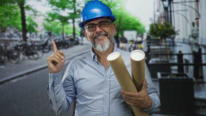 Architect man points finger up and holds rolled plans on street wearing blue hard hat; professional...