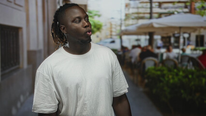 Man purses lips and clasps hands on a street terrace outdoors wearing white t shirt and dreadlocks...