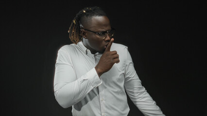 Obraz premium Man young african american shushing with finger to lips wearing white shirt and glasses in studio against black backdrop; silence secrecy.