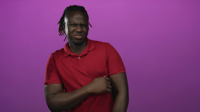 Young african american man grimacing while holding his arm in studio with purple backdrop; disgust reaction.