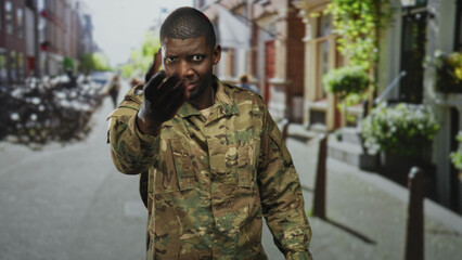 Obraz premium Man soldier in camouflage uniform beckons with hand toward camera, wearing backpack, closeup on a street; pride duty invitation.