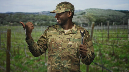 Obraz premium Soldier holding wrench with thumbs up gesture in forest setting among vineyard rows wearing camouflage uniform and cap; pride duty.