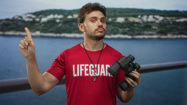 Lifeguard man points finger upward while holding binoculars on seaside railing; public safety vigilance.