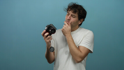 Hispanic man holds camera while pondering idea against vivid blue background, showcasing a...