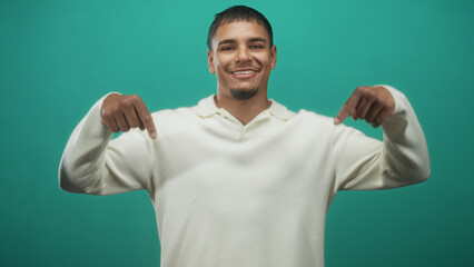 Fototapeta premium Young hispanic man smiling points both index fingers downward at front of white pullover sweater in studio with teal backdrop; friendly confidence.