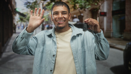 Obraz premium Man holding car keys and waving open hand in a narrow pedestrian street with parked cars and storefronts, smiling broadly at camera; celebration new ownership.