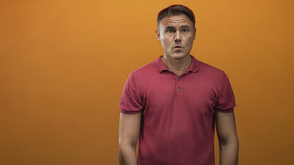 Man covering mouth with hands in a studio against an orange backdrop wearing a red polo shirt and...