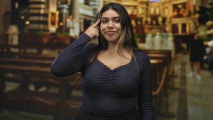 Fototapeta premium Woman pointing to temple while touching hair and smiling with closed eyes, wearing navy top and nose ring inside building; serenity confidence.