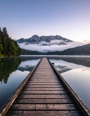 Muelle de madera que se adentra en un lago tranquilo con niebla y monta&ntilde;as al amanecer, reflejando el paisaje sereno y los &aacute;rboles en el agua en calma.