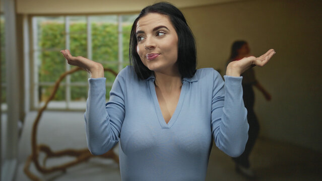 Young hispanic woman stands in an indoor living room gesturing with an expression of indifference and confusion in a relaxed home setting.