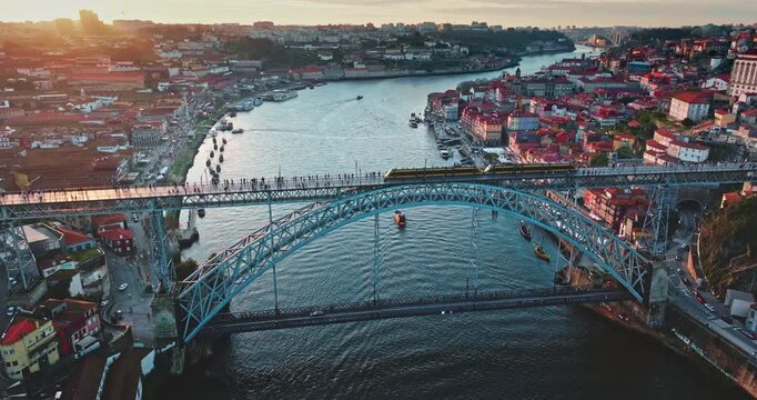Sunset Porto cityscape with Dom Luis I Bridge over the Douro River, historic Ribeira buildings rooftops, waterfront bathed in warm light, boats floating under train riding railway. Aerial drone shot