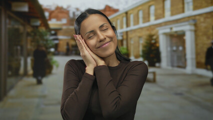Fototapeta premium Young hispanic woman smiling peacefully with closed eyes on a quiet outdoor street with background buildings and trees, embodying serenity and relaxation.