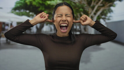 Naklejka premium Young hispanic woman outdoors screaming with eyes closed and fingers in ears, expressing frustration with blurred green trees in the background.