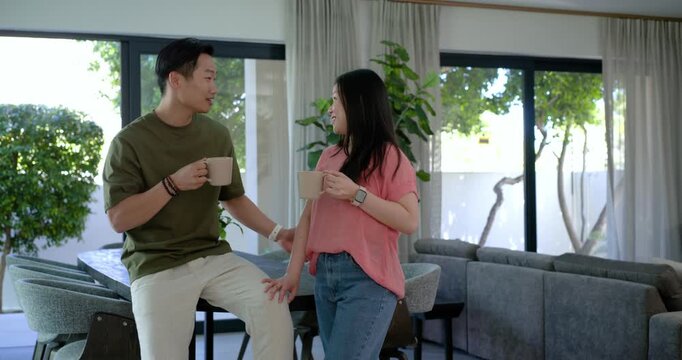 Asian couple sitting and standing at dining counter locking eyes, holding mugs, showing affection