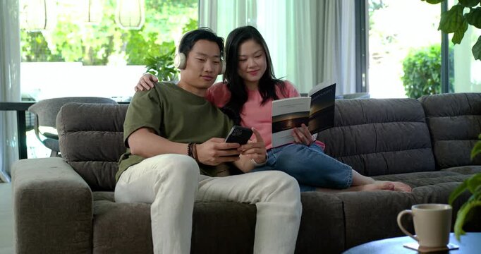 Diverse couple tapping smartphone, leaning on gray sofa, reviewing booklet and laughing