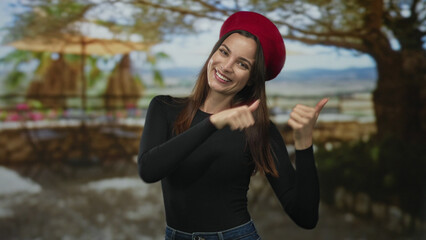 Fototapeta premium Woman points finger with smiling face and visible hands wearing red beret on street terrace; playful charm.