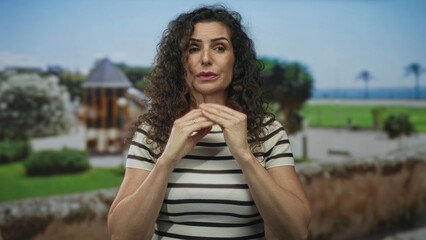 Woman in a striped shirt grips her clasped hands near chest in front of a building wall under bright sunlight; anxiety.