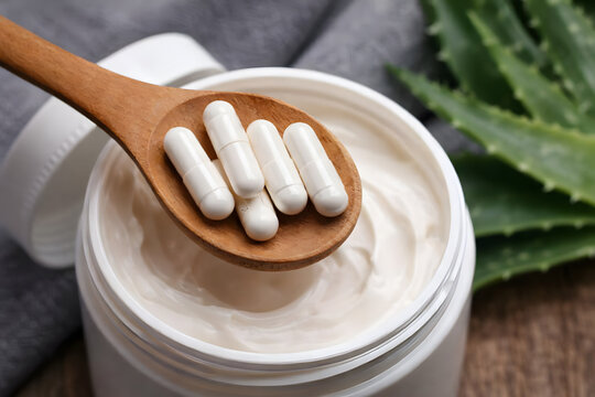 A wooden spoon holds white capsules over a jar of cream with aloe vera in the background
