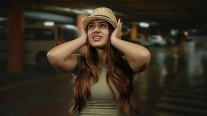 Young woman wearing hat expressing frustration in an indoor parking area with a distressed look amid dim lighting and parked cars. © Krakenimages.com