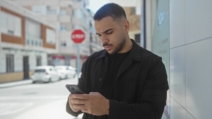 Hispanic man taps smartphone screen on sunlit street against modern building facade; focus...