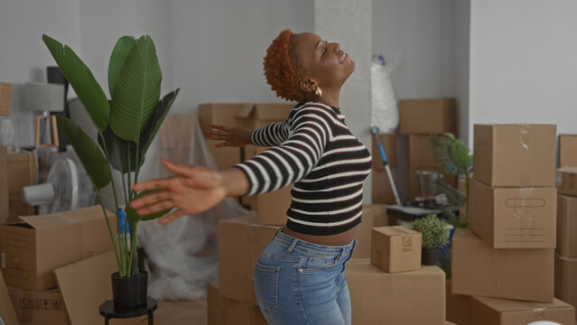Young woman stretches arms and smiles broadly amid stacked cardboard boxes in a spacious building; optimism.