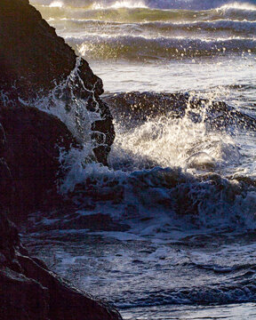 View of powerful, foamy waves crashing against dark, rugged rocks, the ocean spray illuminated by sunlight, creating a dynamic coastal scene, Fort Bragg, California, United States.